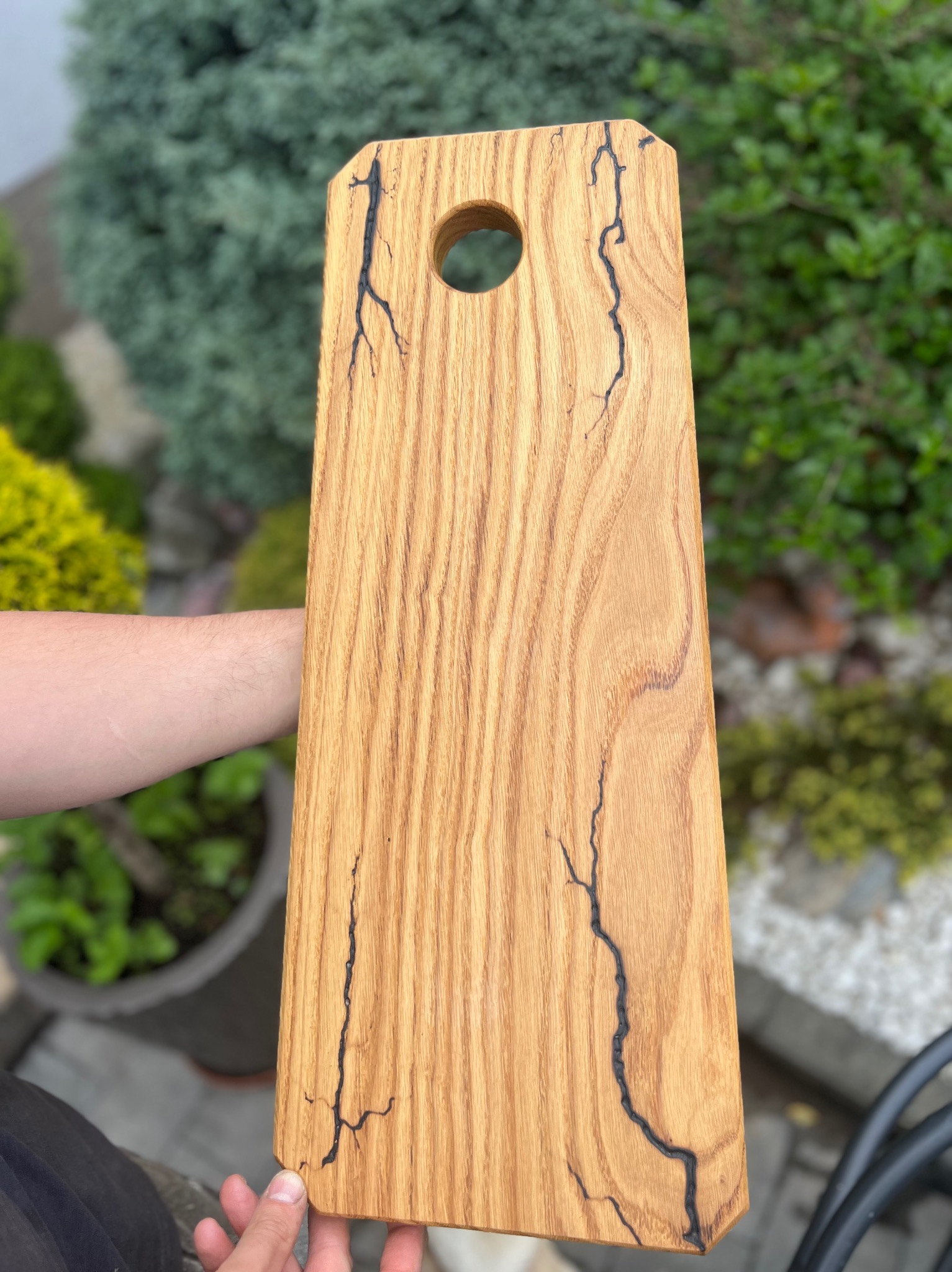 Deska do krojenia z jasnego drewna dębowego z unikalnymi czarnymi wzorami przypominającymi pioruny, trzymana w dłoni na tle zieleni ogrodowej.