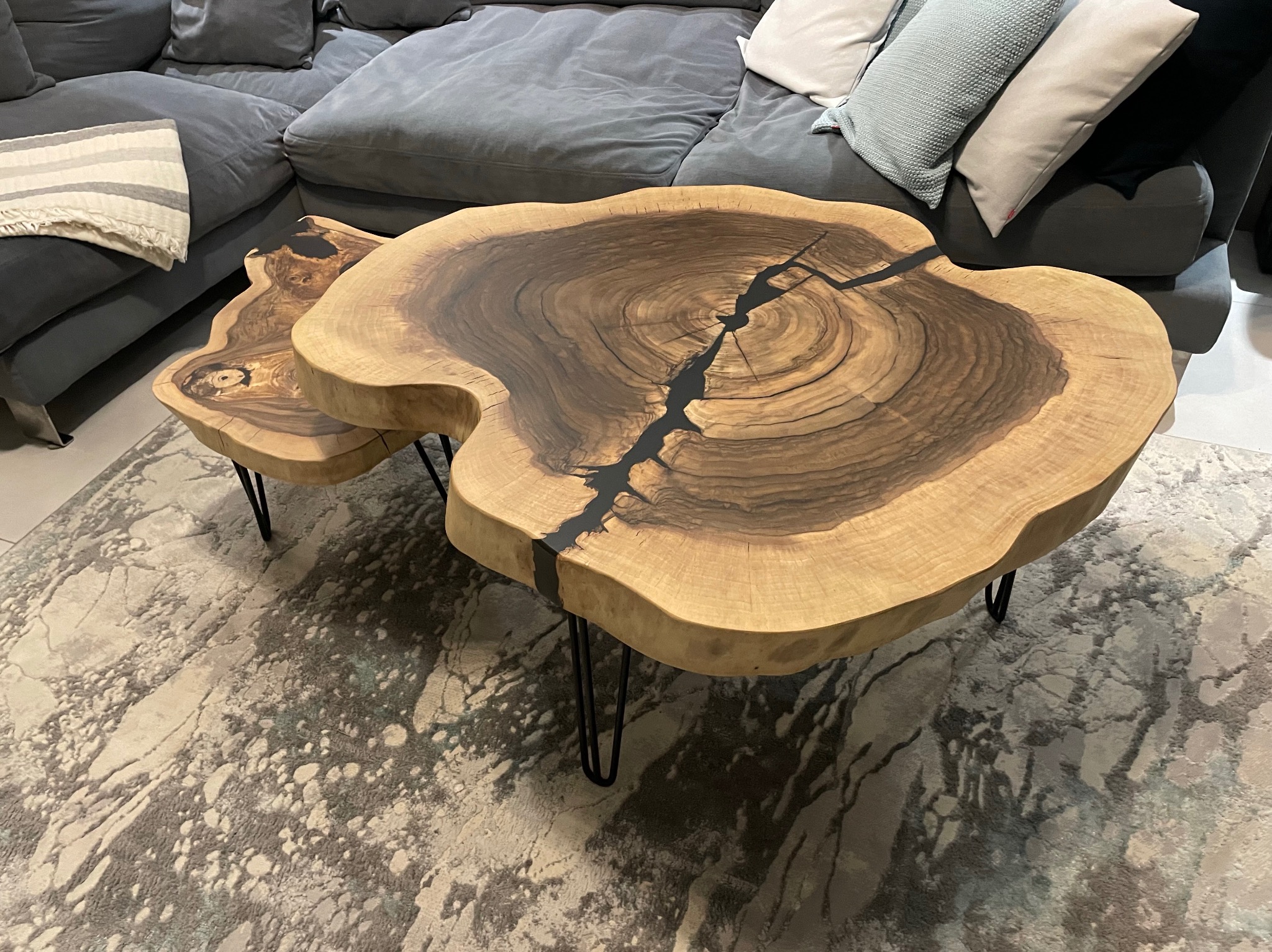 Dwa stoliki kawowe z blatem z litego drewna o nieregularnym kształcie, wypełnionym czarną żywicą, na cienkich, metalowych nóżkach, ustawione na wzorzystym dywanie w salonie z szarą sofą w tle.