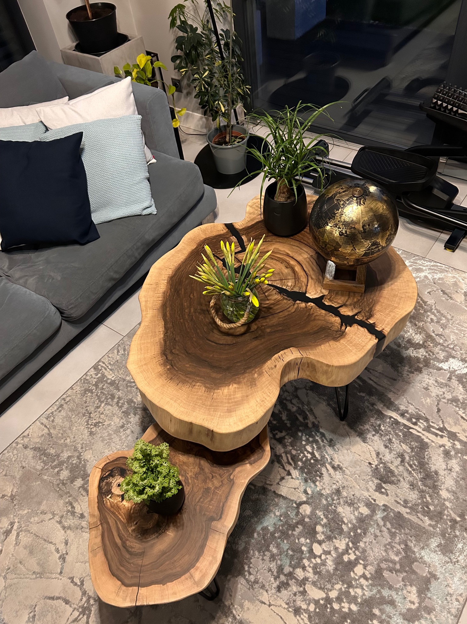 Dwupoziomowy stolik kawowy z plastra drewna z żywicą epoksydową, na metalowych nóżkach, z dekoracjami roślinnymi i globusem, na wzorzystym dywanie w salonie.