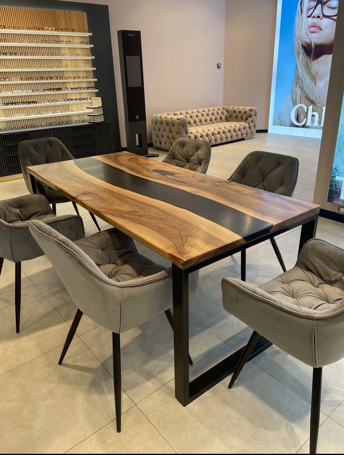 Stół z blatem z drewna orzechowego z żywicą epoksydową, otoczony sześcioma szarymi krzesłami z tapicerką, w tle ekspozycja okularów i beżowa sofa.