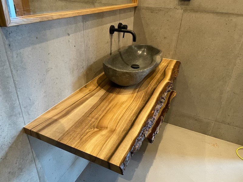 Drewniany blat łazienkowy z naturalną krawędzią kory, na którym umieszczona jest kamienna umywalka w kształcie misy. Minimalistyczna czarna bateria ścienna. Betonowe płytki na ścianie i podłodze.