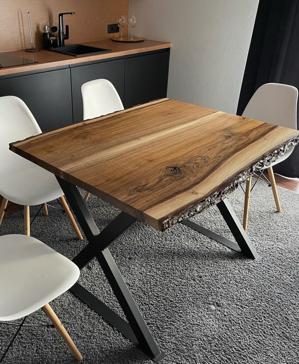 Kwadratowy stół z blatem z litego drewna orzechowego o naturalnej krawędzi, wsparty na czarnych, metalowych nogach w kształcie litery X, otoczony białymi krzesłami w nowoczesnej kuchni z czarnymi...