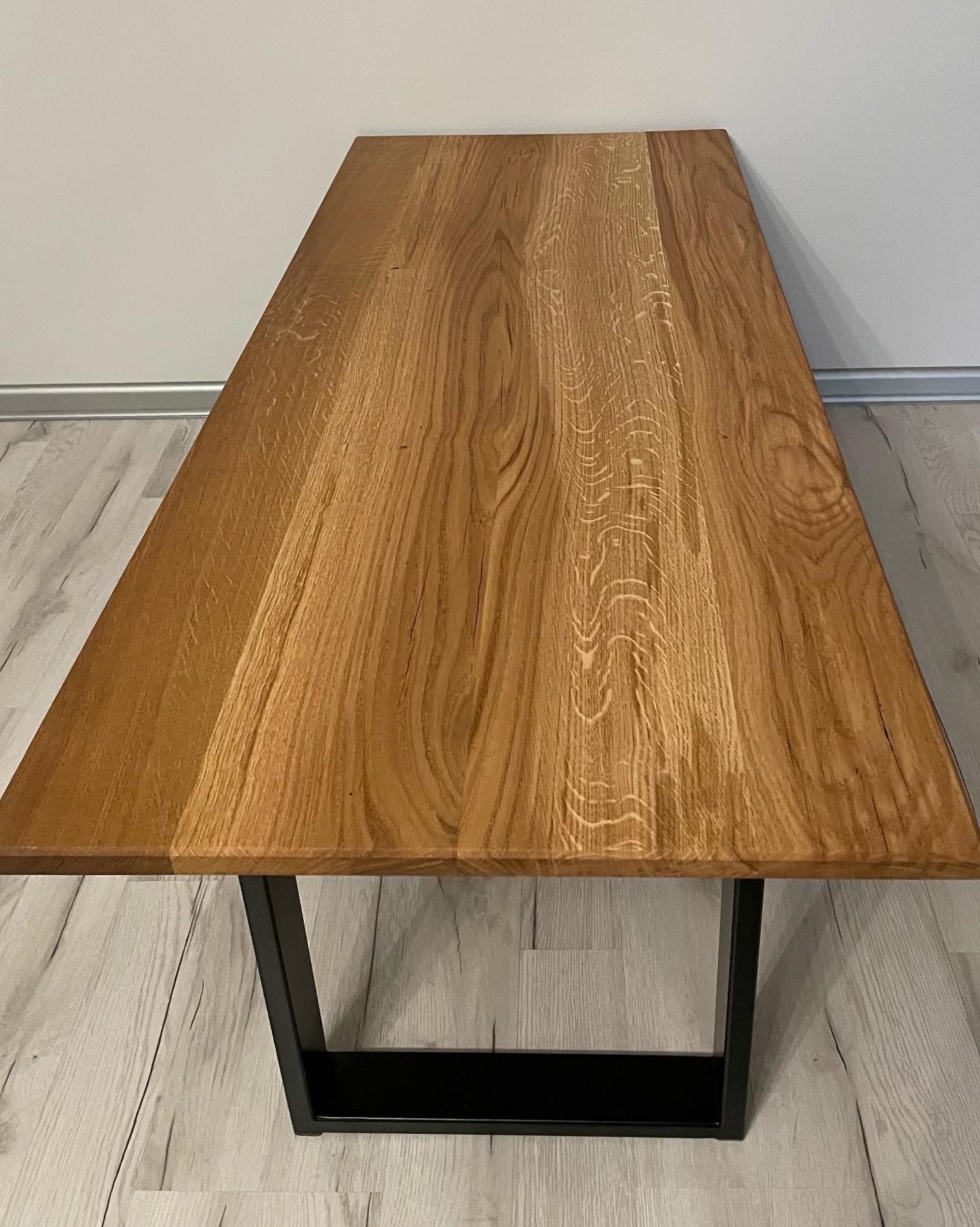 Prostokątny blat stołu z jasnego drewna o wyraźnym usłojeniu, osadzony na czarnych, metalowych nogach w kształcie litery V, na tle szarej ściany i podłogi imitującej drewno.