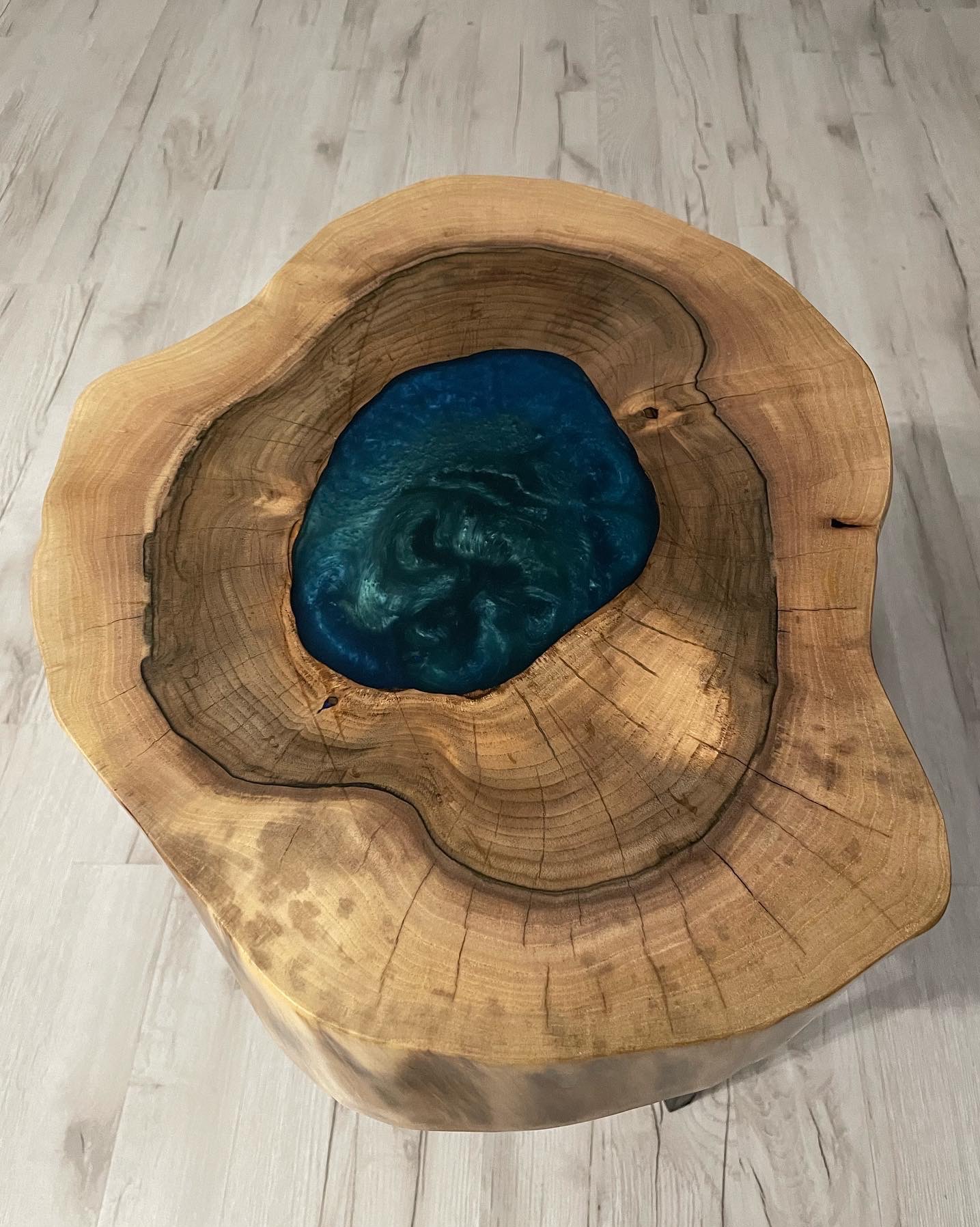 Stolik kawowy z blatu z litego drewna z żywicą epoksydową w kolorze turkusowym, ukazujący słoje drzewa i naturalne pęknięcia, na tle paneli podłogowych imitujących drewno.