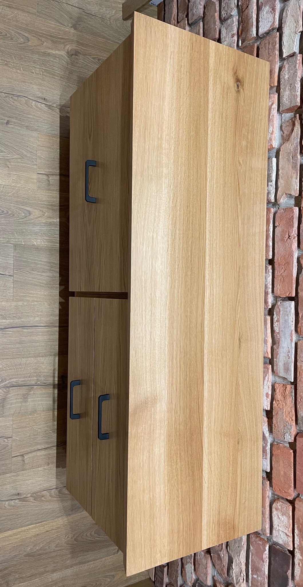 Drewniana szafka wisząca z dwiema parami drzwi z czarnymi uchwytami, zamontowana na tle ściany z czerwonej cegły i drewnianej podłogi.