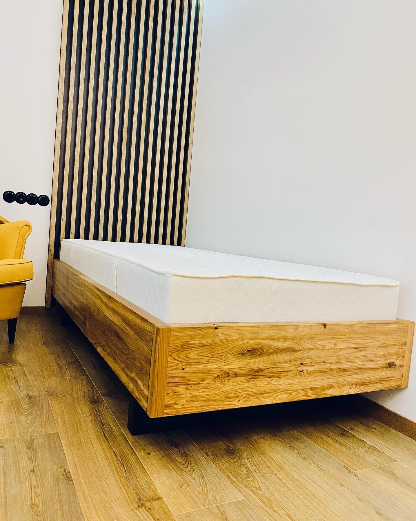 Drewniane łóżko z materacem w minimalistycznej sypialni z drewnianą ścianką działową i żółtym fotelem w tle.