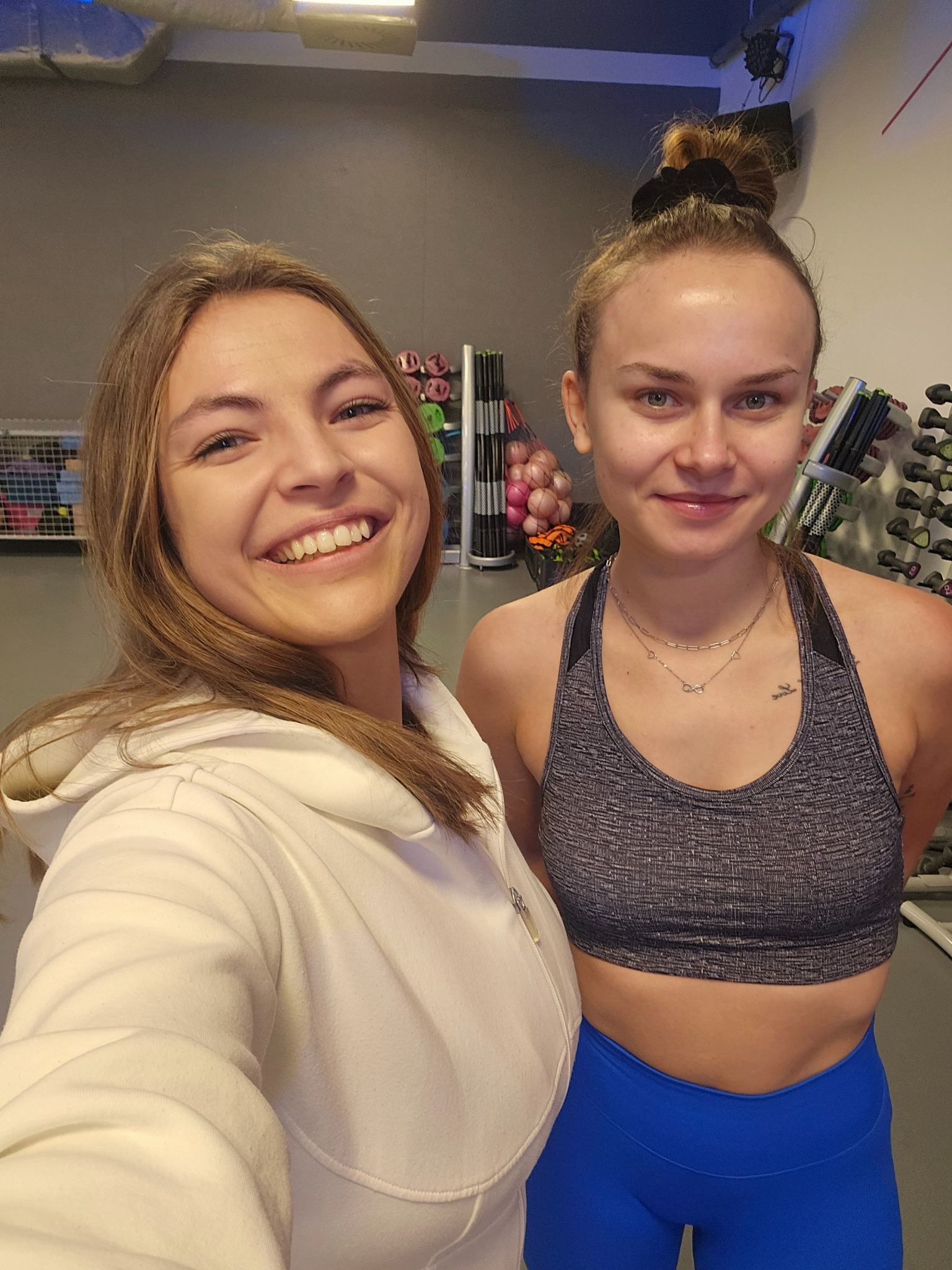 Dwie uśmiechnięte kobiety w strojach sportowych pozują w siłowni, widoczne hantle i sprzęt do ćwiczeń w tle.