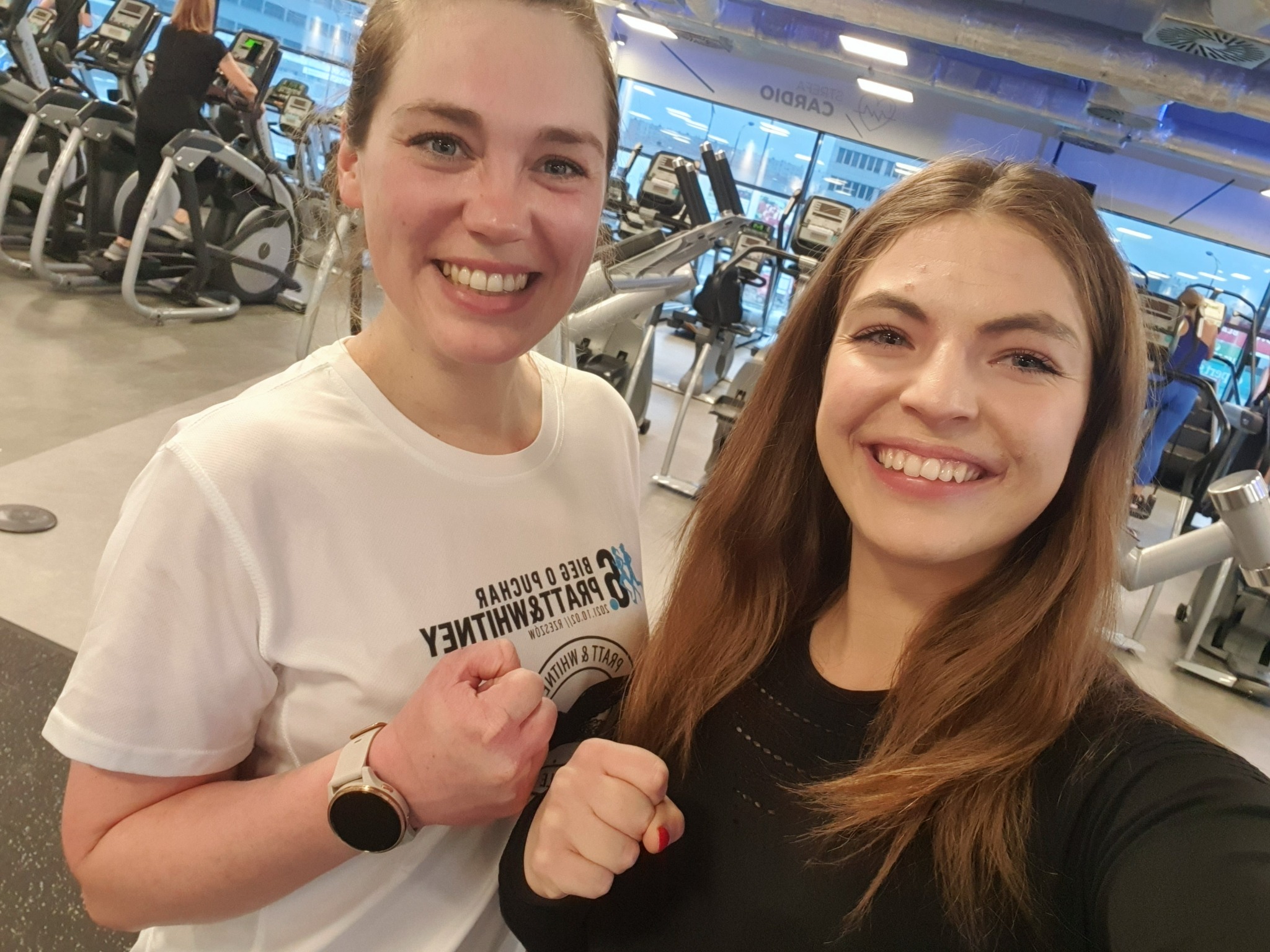Dwie uśmiechnięte kobiety w siłowni, z zaciśniętymi pięściami, świętują udany trening na tle sprzętu cardio i okien z widokiem na miasto.