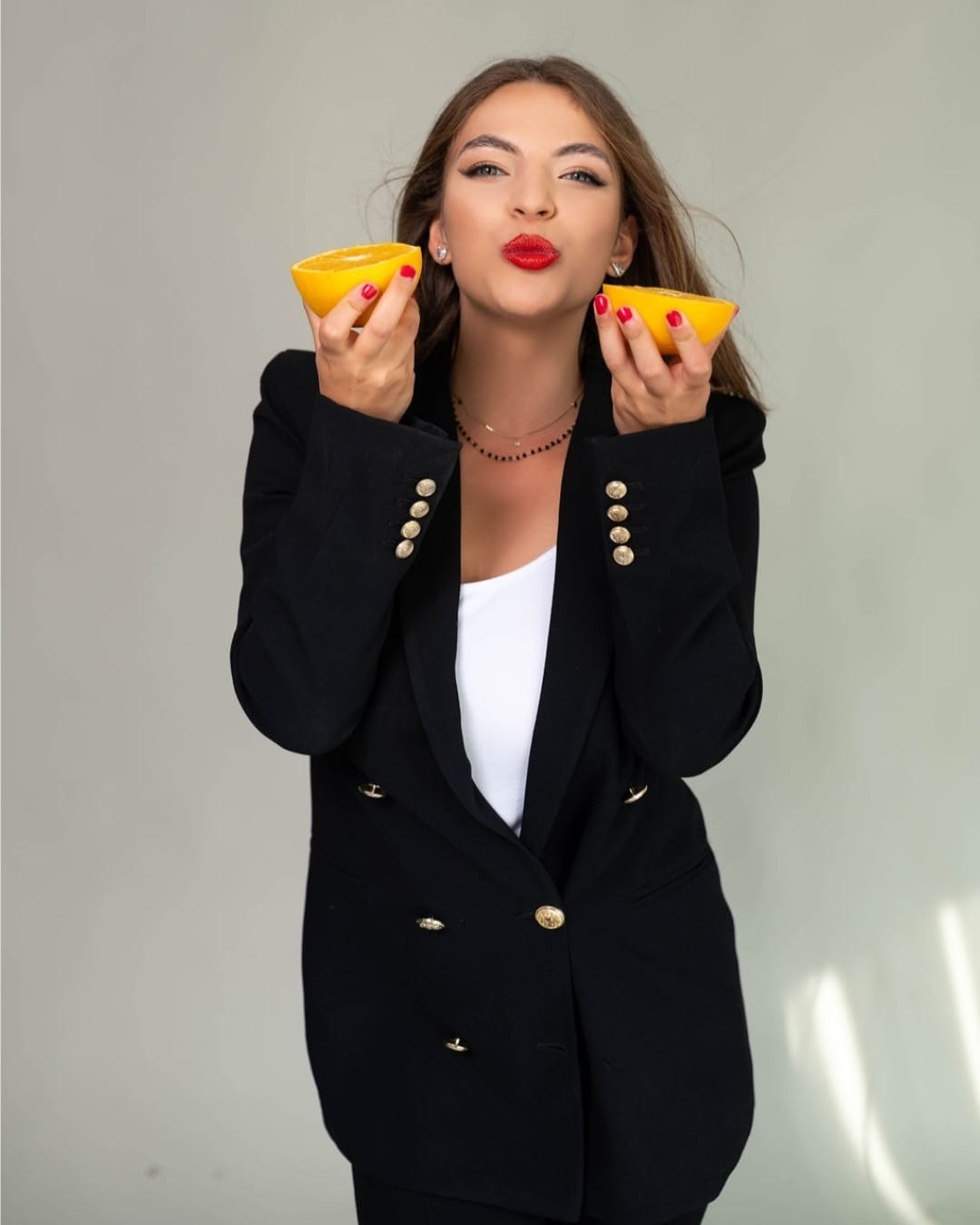 Kobieta w czarnej marynarce z pomarańczowymi paznokciami trzyma dwie połówki pomarańczy, usta pomalowane na czerwono, lekkie zmarszczki mimiczne wokół oczu.