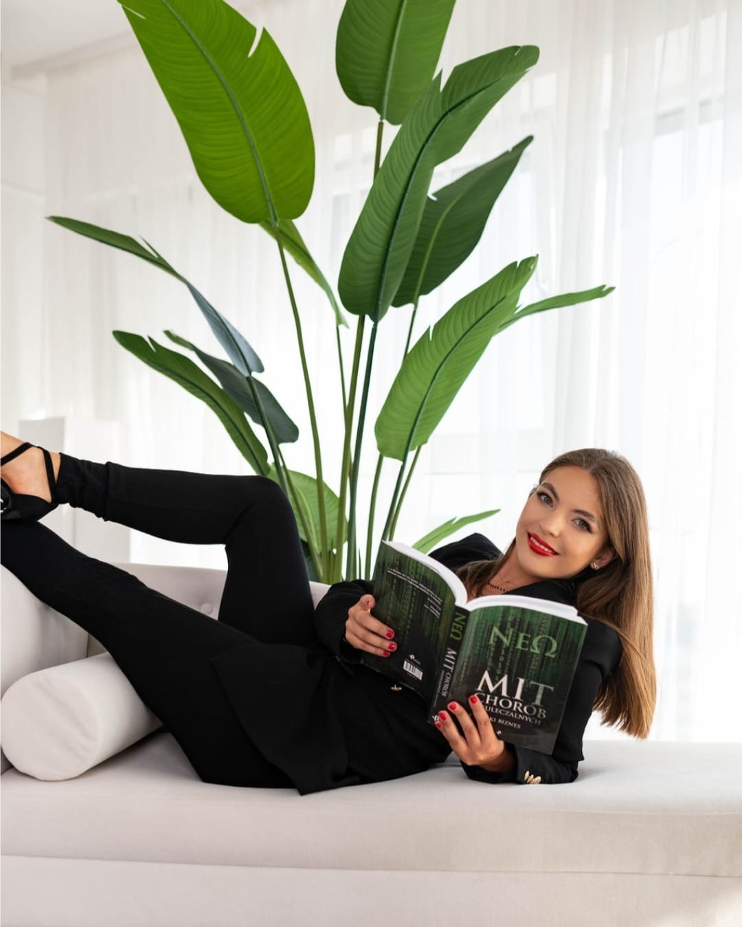 Kobieta w czarnym garniturze leży na białej sofie, czytając książkę o chorobach uleczalnych biznesu, z rośliną w tle.