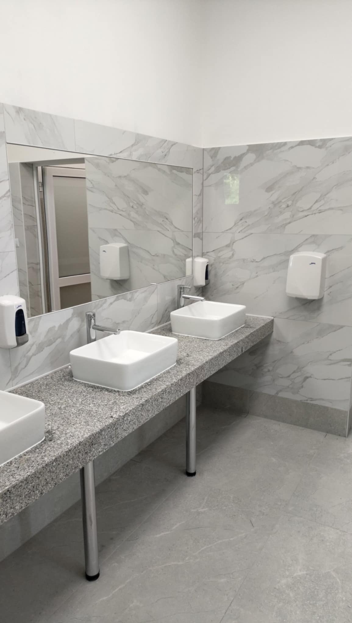 Umywalki nablatowe w publicznej toalecie, blaty z szarego lastryko, ściany wyłożone marmurowymi płytkami, srebrne baterie i dozowniki mydła.
