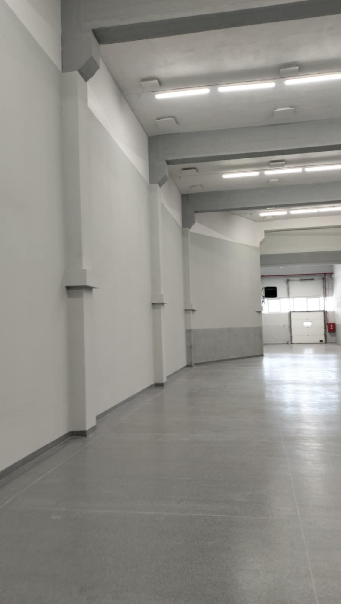 Wnętrze hali magazynowej z szarymi ścianami, betonową podłogą i widocznymi elementami konstrukcyjnymi pod sufitem, oświetlone lampami jarzeniowymi, z otwartą bramą w tle.