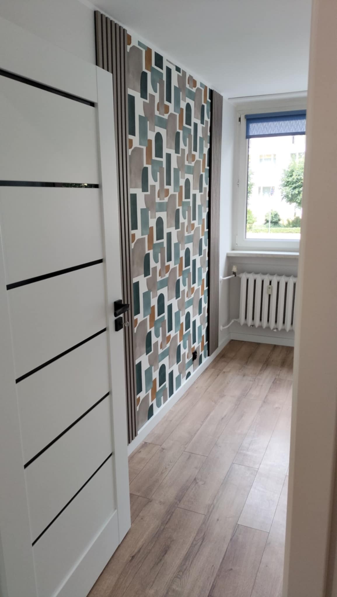 Wnętrze pokoju z panelami ściennymi z abstrakcyjnym wzorem, drewnianymi listwami dekoracyjnymi, białymi drzwiami z czarnymi poziomymi paskami i grzejnikiem pod oknem.