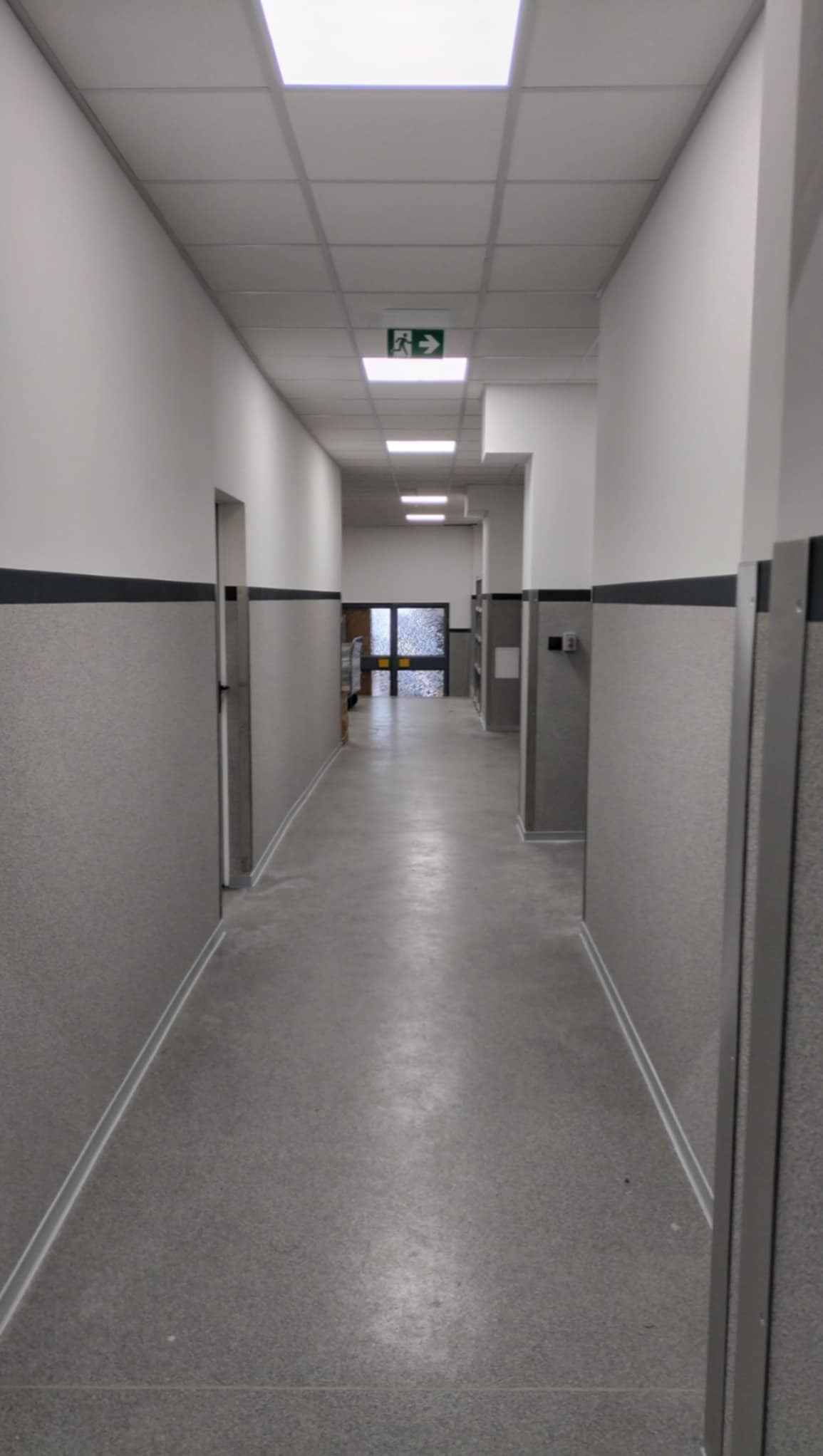 Długi korytarz w budynku użyteczności publicznej z szarymi panelami na ścianach do połowy wysokości, białymi ścianami powyżej i szarą podłogą; widoczny znak ewakuacyjny na suficie.