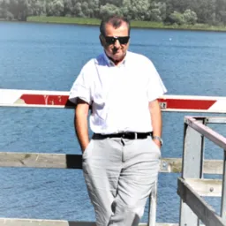 Mężczyzna w okularach przeciwsłonecznych, białej koszuli i szarych spodniach, oparty o barierkę z widokiem na jezioro i las w tle.