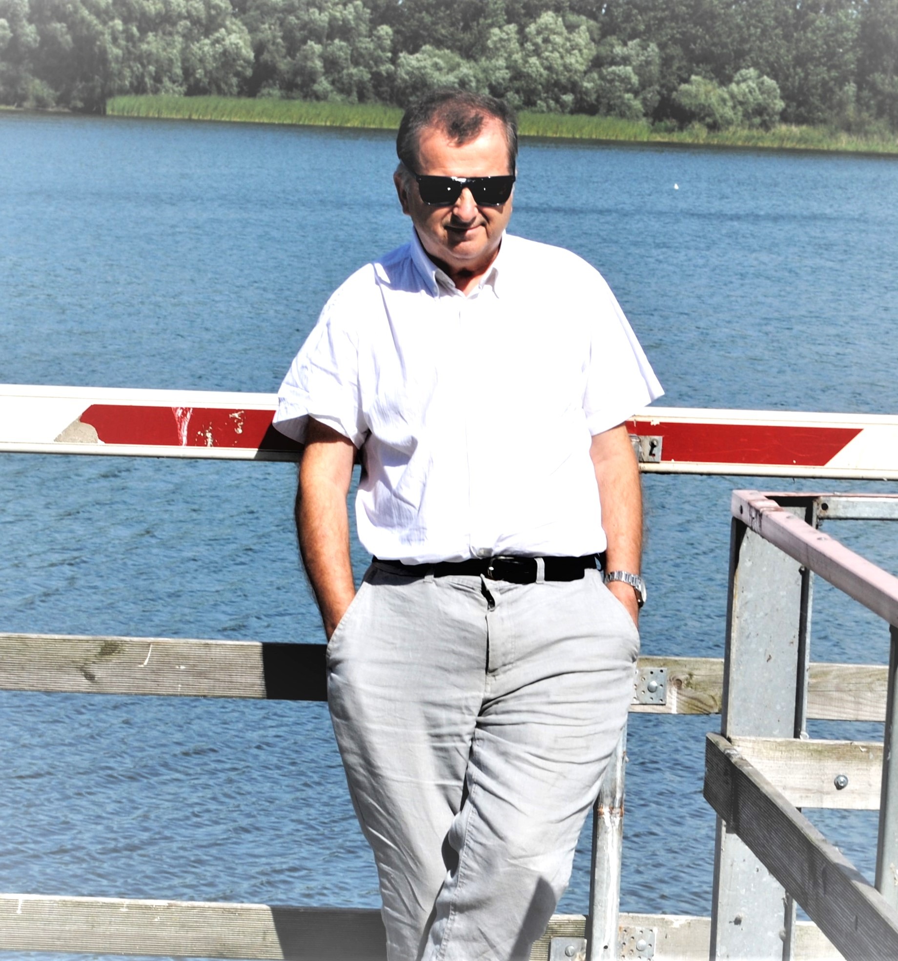 Mężczyzna w okularach przeciwsłonecznych, białej koszuli i szarych spodniach, oparty o barierkę z widokiem na jezioro i las w tle.