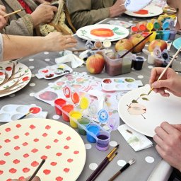 Izabela Kot kotydoroboty - Grupa osób maluje ceramiczne talerze farbami akrylowymi, używając pędzli i palet kolorów, na stole w kropki, z jabłkami i wzornikami.
