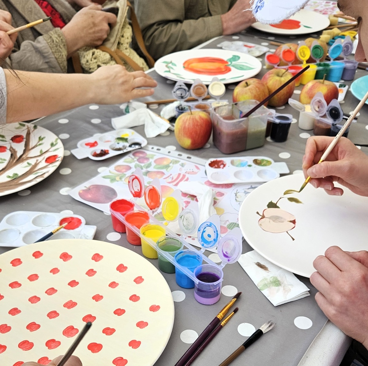Grupa osób maluje ceramiczne talerze farbami akrylowymi, używając pędzli i palet kolorów, na stole w kropki, z jabłkami i wzornikami.