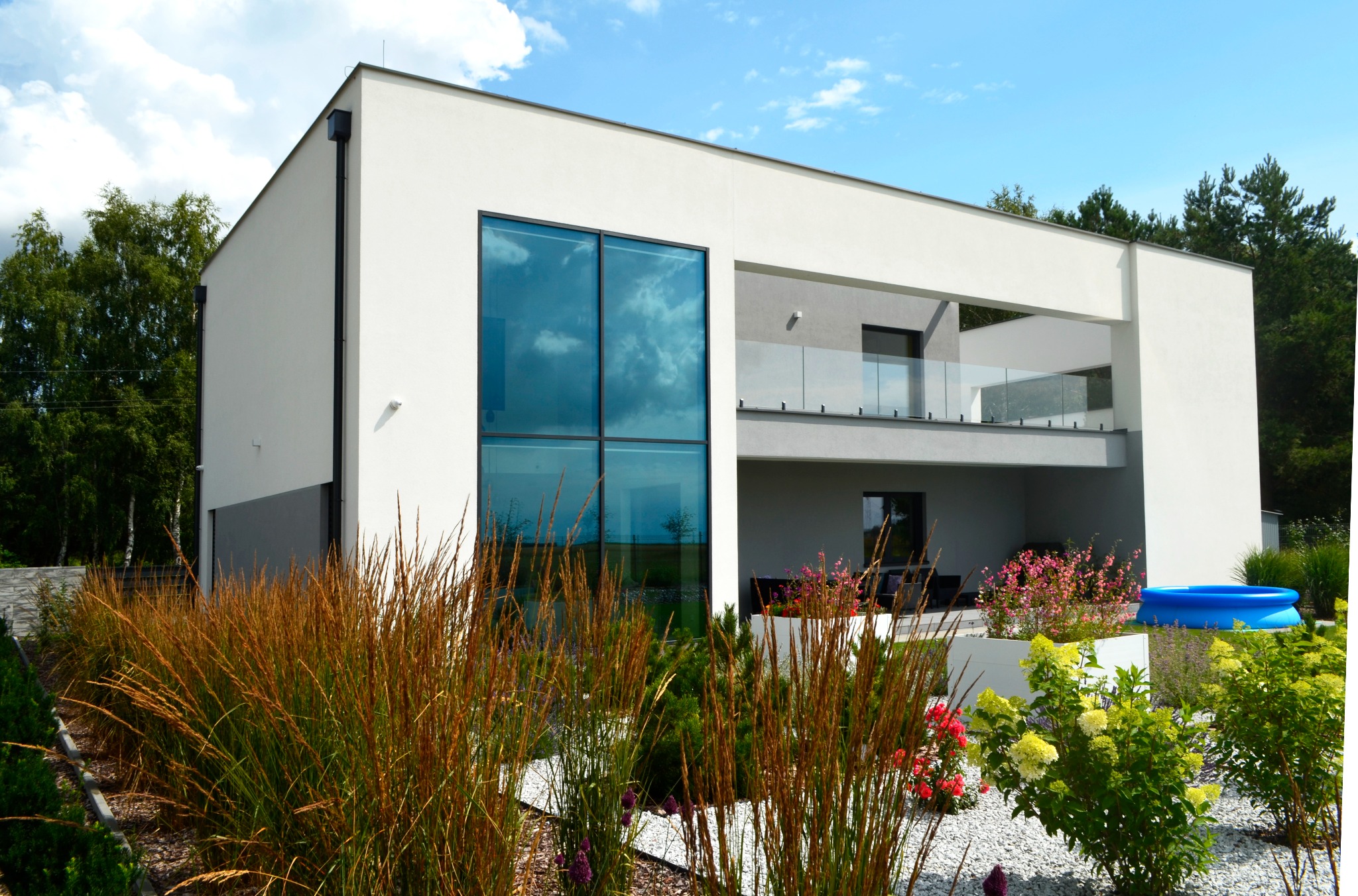 Nowoczesny dom jednorodzinny z dużymi przeszkleniami, tarasem z balustradą ze szkła i ozdobnym ogrodem z trawami i kwiatami. W tle widoczne drzewa i błękitne niebo.