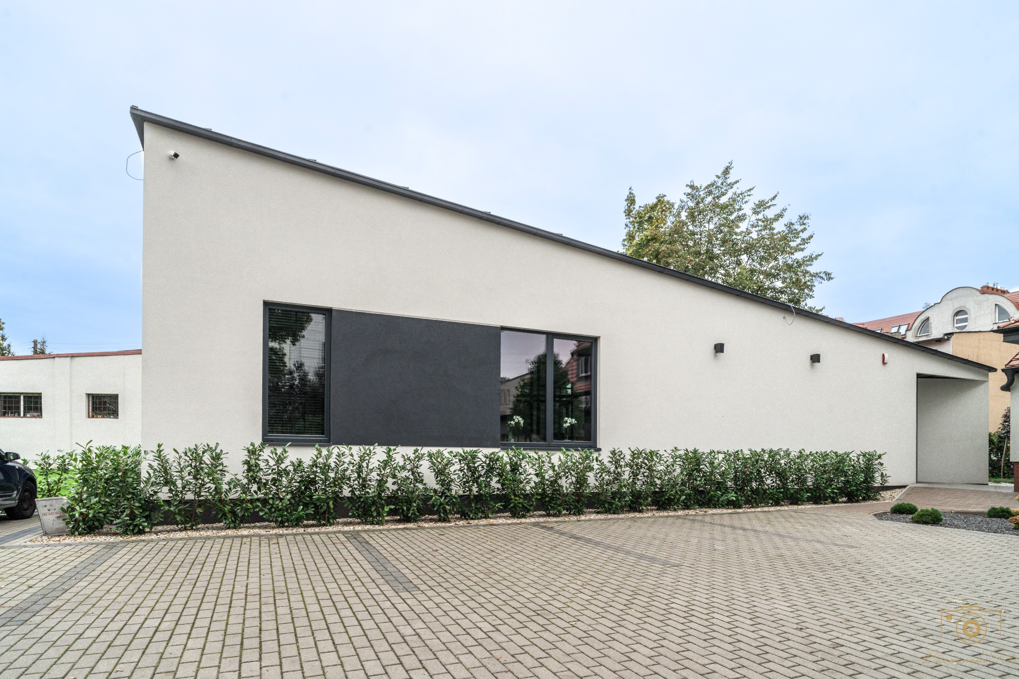 Nowoczesny dom jednorodzinny z płaskim dachem i dużymi oknami, minimalistyczna elewacja w odcieniach bieli i szarości, otoczony niskimi krzewami i brukowanym podjazdem.