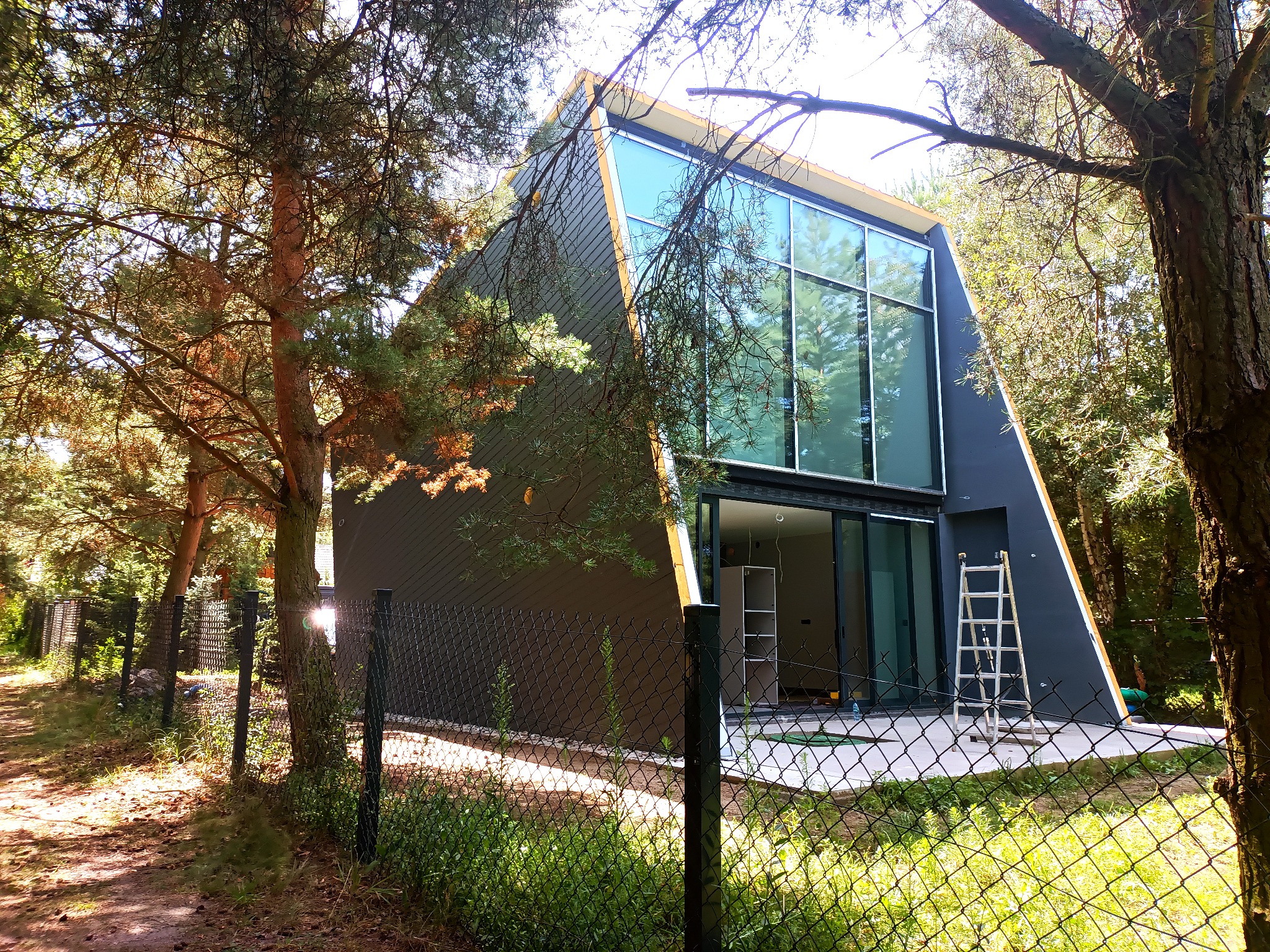 Nowoczesny dom w kształcie trójkąta z dużymi przeszkleniami, widziany zza ogrodzenia, z drabiną opartą o ścianę i widocznym wnętrzem w trakcie wykończenia.