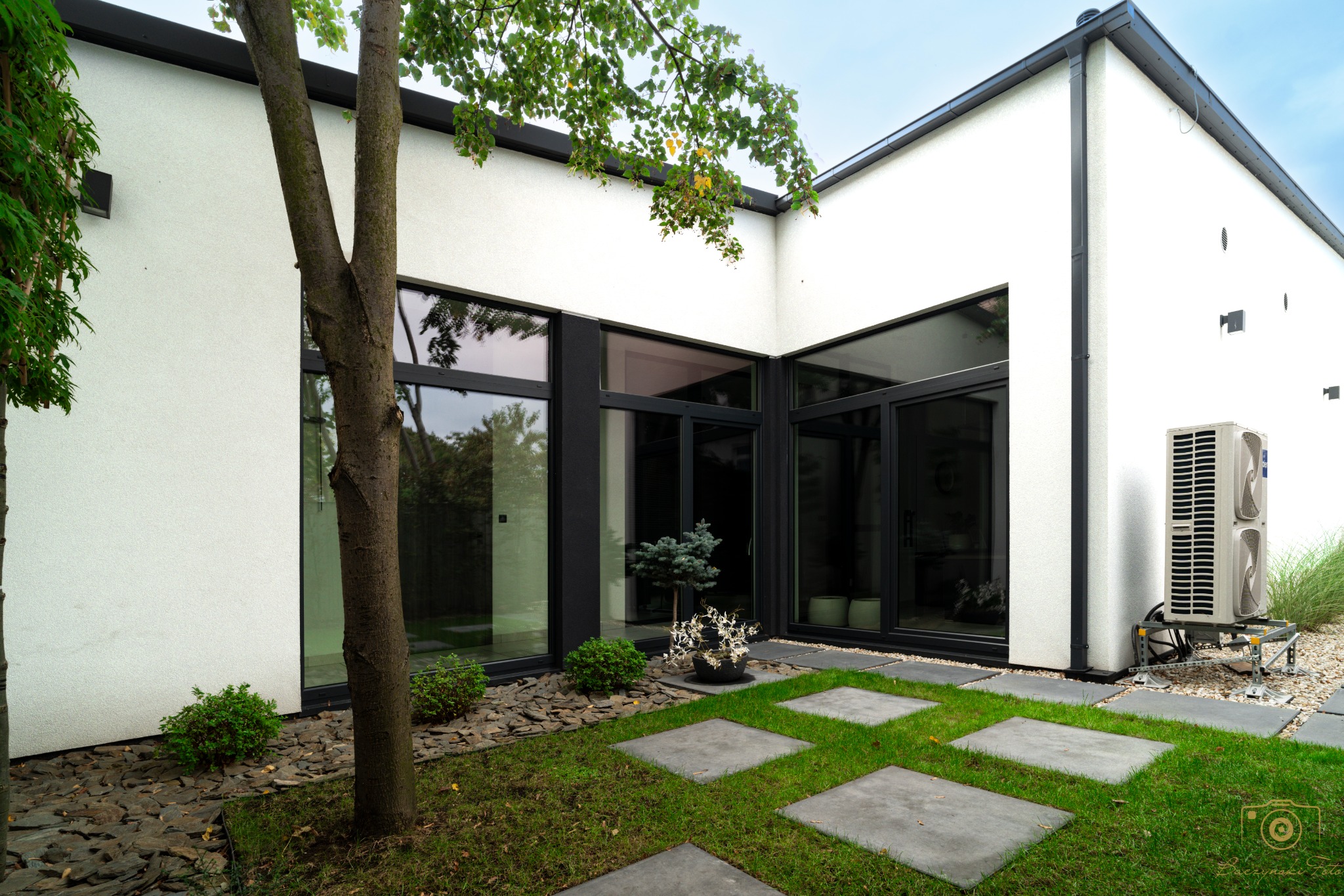 Nowoczesny dom z białymi ścianami i czarnymi ramami okiennymi, otoczony zielonym trawnikiem z betonowymi płytami, drzewami i krzewami, z widoczną jednostką zewnętrzną klimatyzacji na metalowym...