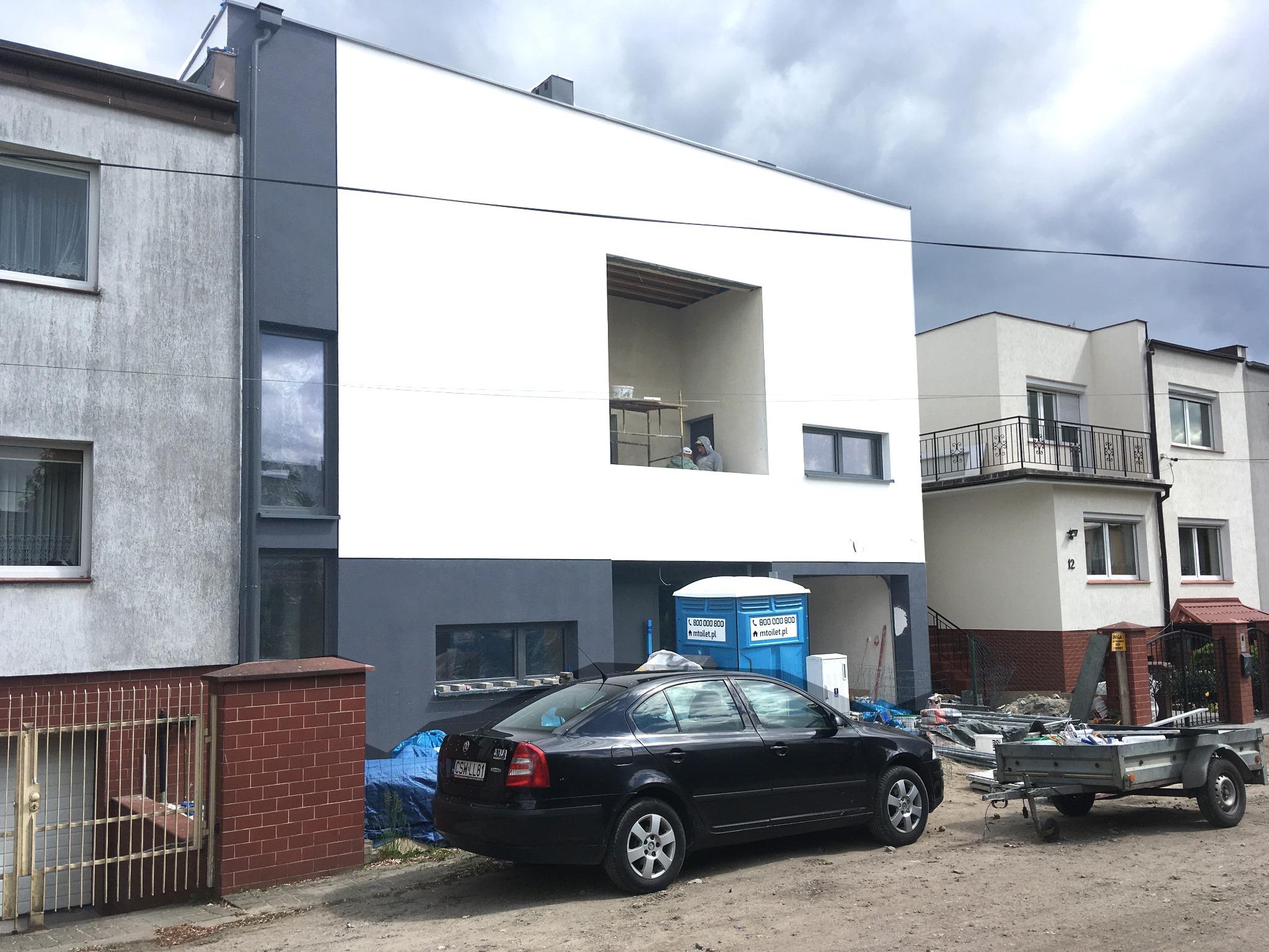 Nowoczesny dom w trakcie budowy z wyraźnie zaznaczonym oknem bez szyby, z pracownikiem w środku, zaparkowany samochód osobowy i przyczepa na zewnątrz, przenośna toaleta.