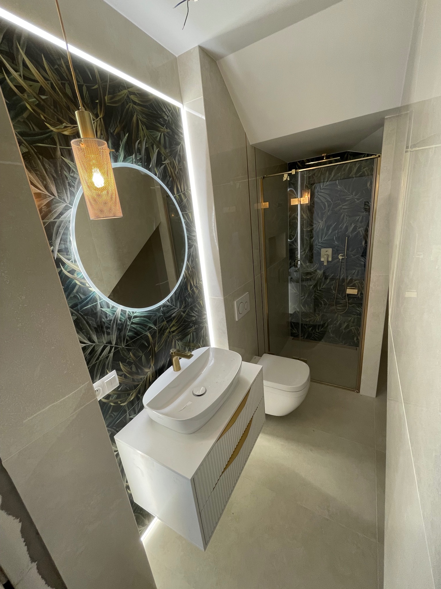 Nowoczesna łazienka z tropikalną tapetą, okrągłym lustrem podświetlanym LED, białą szafką z umywalką i kabiną prysznicową ze złotymi detalami.