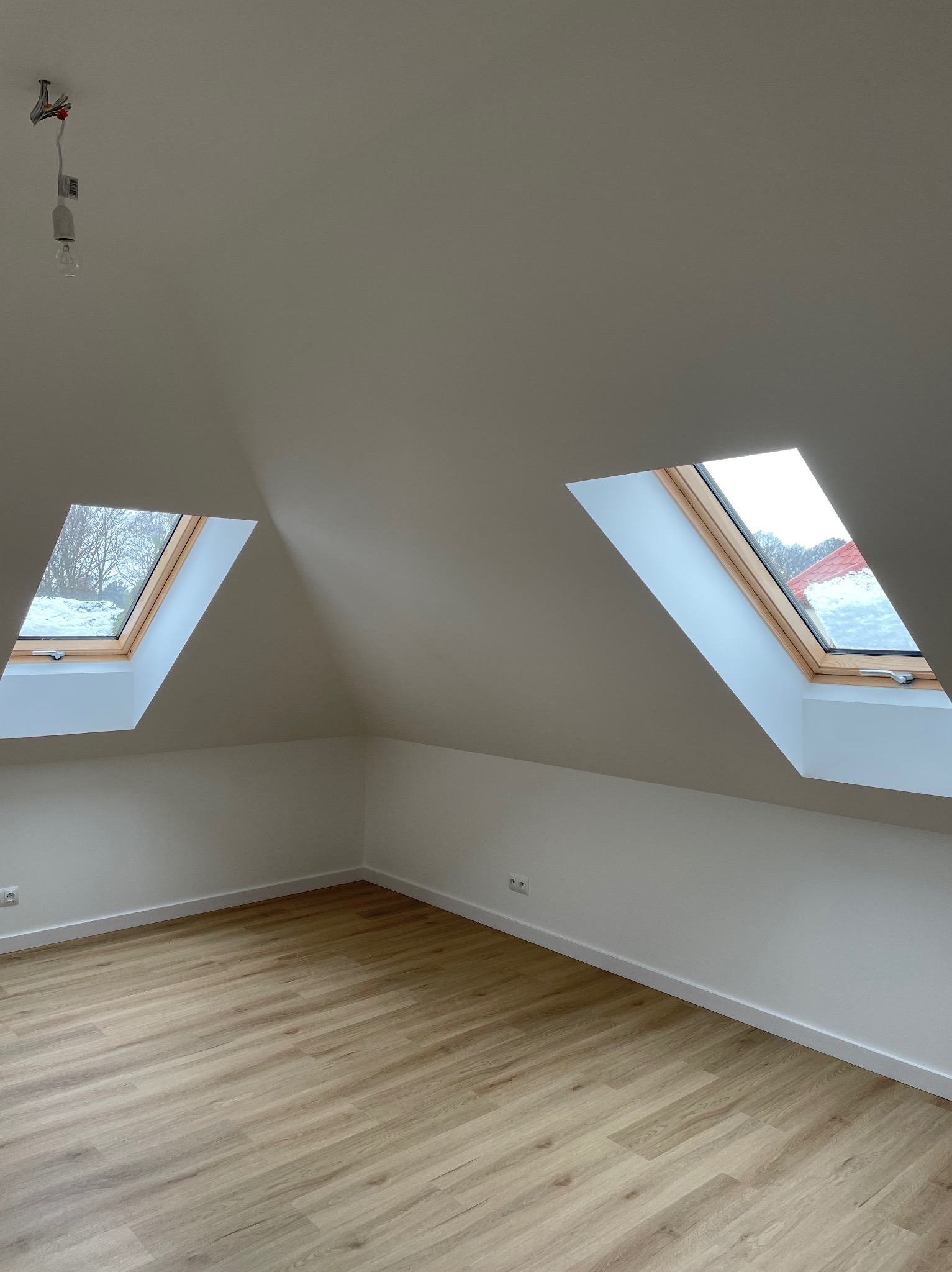 Poddasze z dwoma oknami dachowymi, wykończone drewnianą podłogą. Widoczne gniazdka elektryczne i wisząca żarówka.