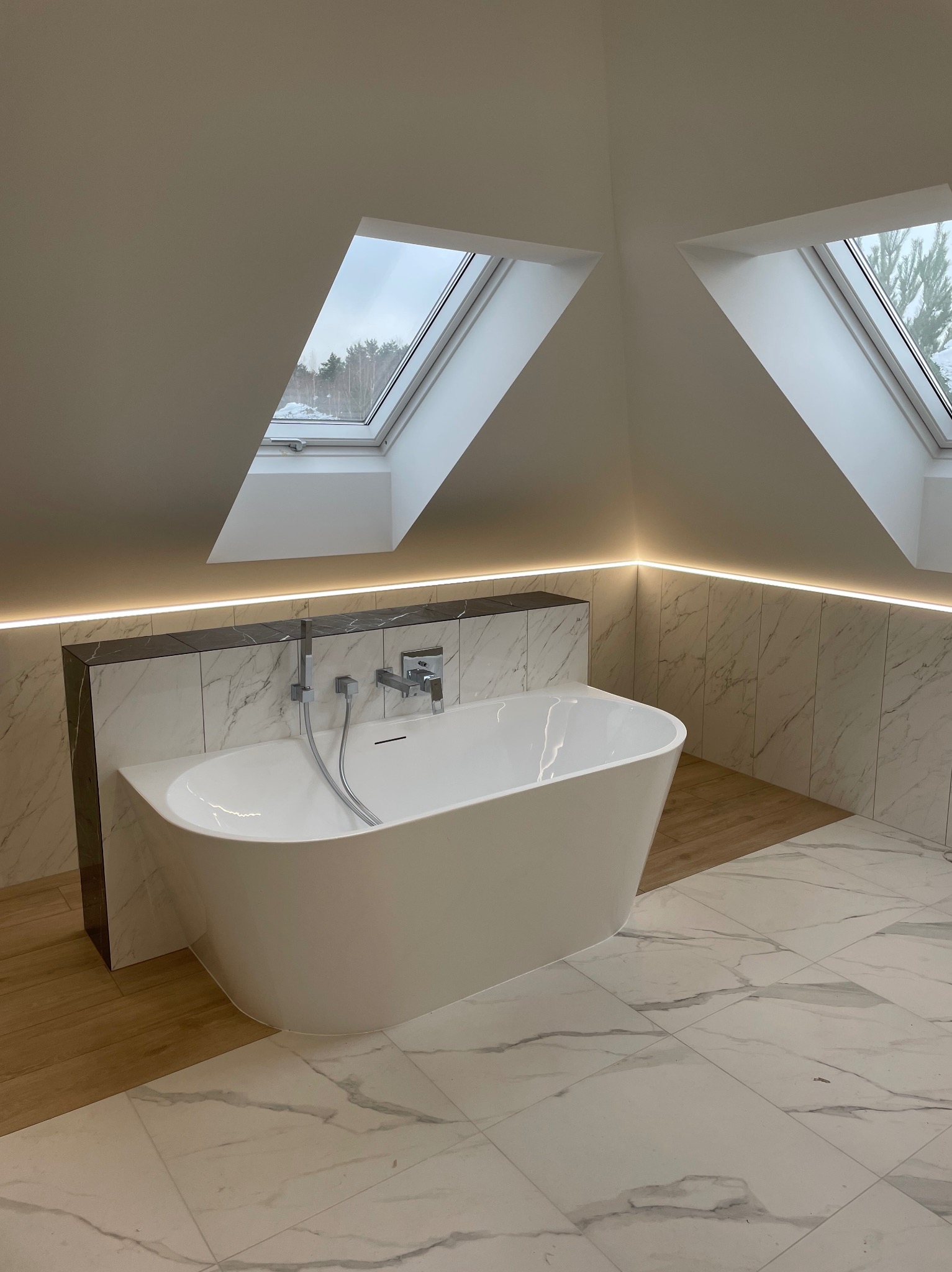 Nowoczesna, biała wanna wolnostojąca z chromowaną baterią, umieszczona w jasnej łazience na poddaszu z marmurową okładziną i dwoma oknami dachowymi z widokiem na zimowy krajobraz.