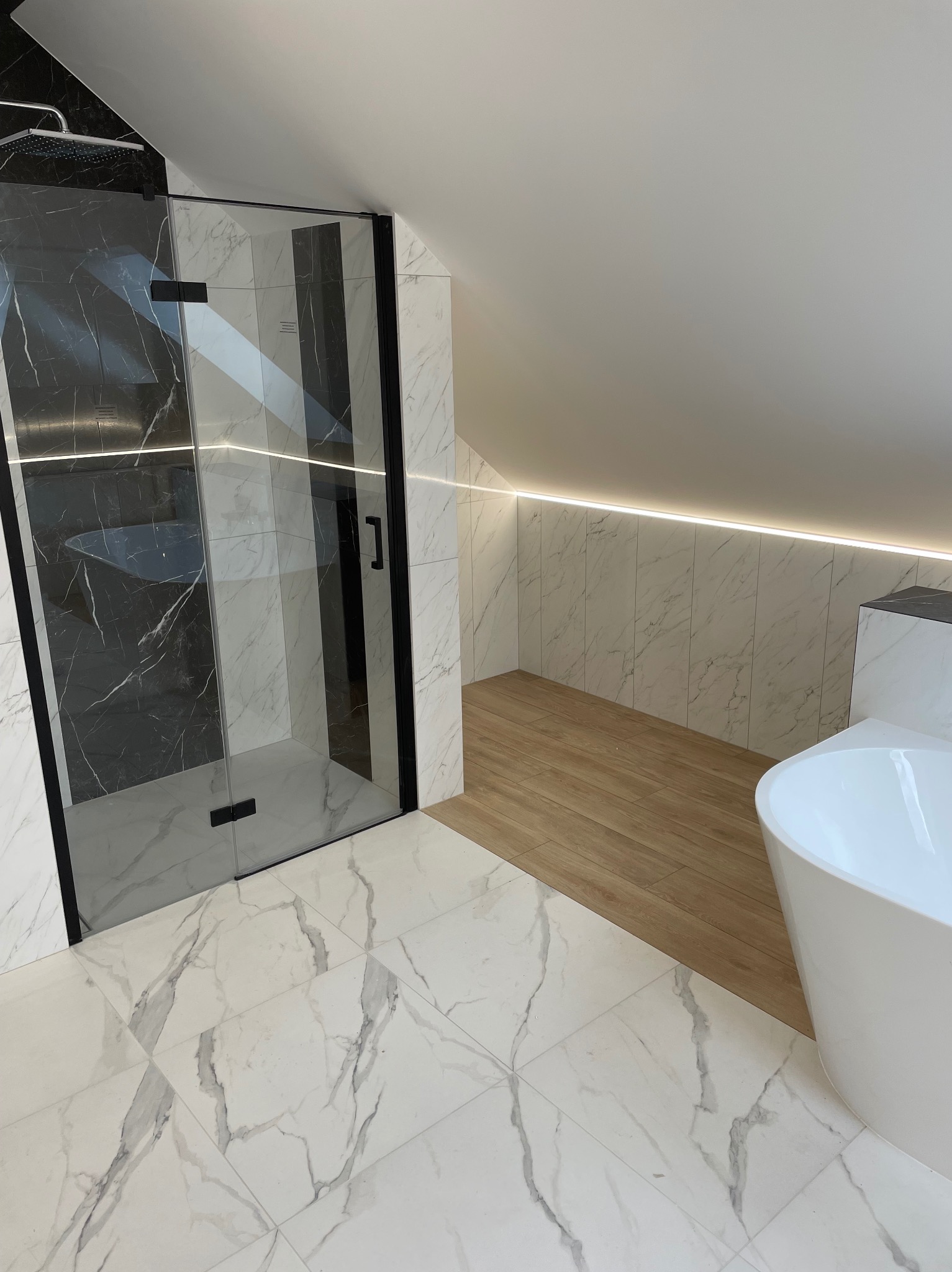 Nowoczesna łazienka na poddaszu z kabiną prysznicową typu walk-in z czarnymi ramami, białą wanną wolnostojącą i marmurowymi płytkami w dwóch kolorach oraz oświetleniem LED.
