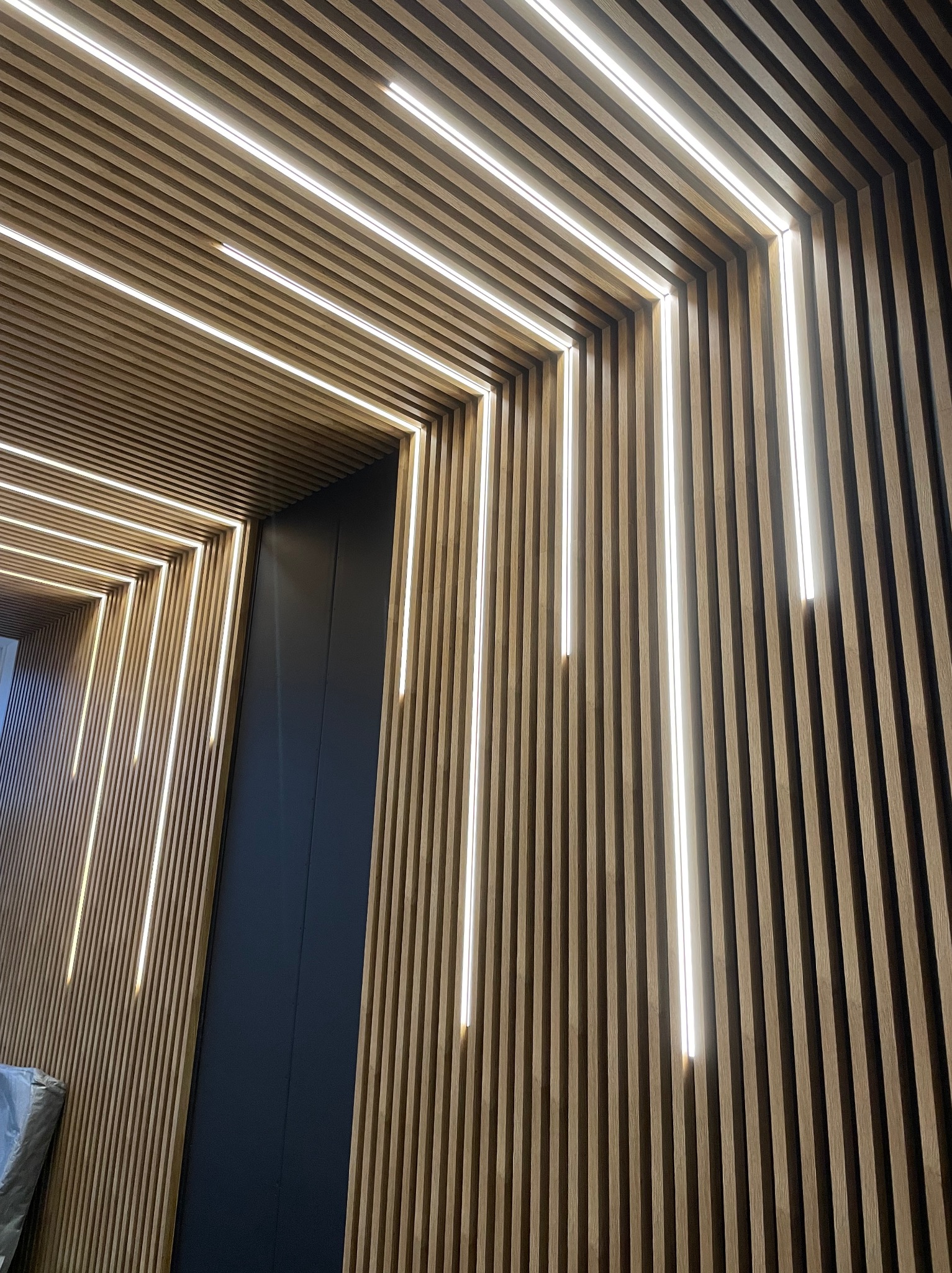 Drewniane lamele na ścianie i suficie z wbudowanym oświetleniem LED tworzące geometryczny wzór.