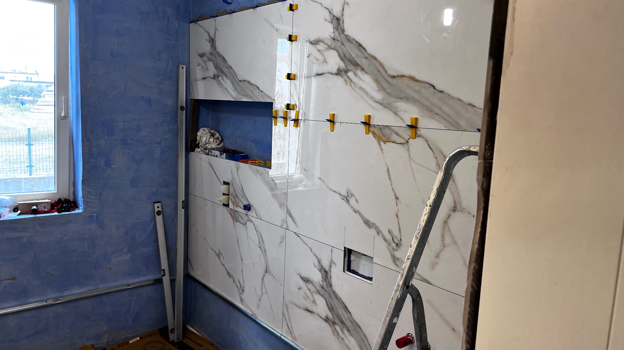 Ściana w trakcie układania płytek imitujących marmur, z widocznymi krzyżykami dystansowymi i poziomnicą, w tle surowa, niebieska ściana i drabina.