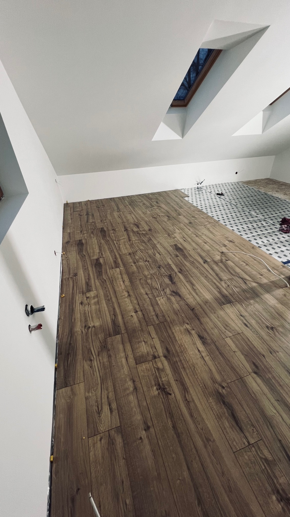 Układanie paneli podłogowych o wzorze drewna w pokoju na poddaszu, widoczne okno dachowe i fragment przygotowanego podłoża pod płytki.