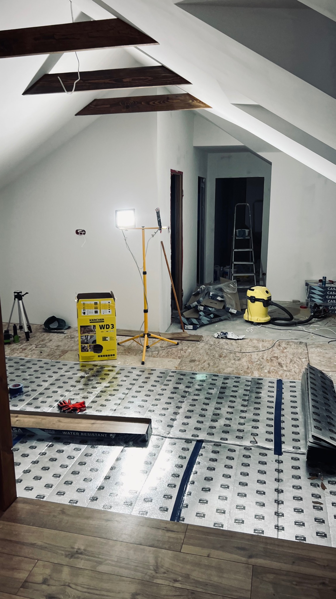 Montaż podłogi: widok na podkład izolacyjny z nadrukiem, lampę budowlaną, odkurzacz przemysłowy i narzędzia w surowym wnętrzu poddasza z drewnianymi belkami.