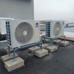 MAT-CAR Mateusz Woźniak - Dwa klimatyzatory Gree zamontowane na dachu budynku, na betonowych podstawach z metalowymi wspornikami. Widoczne budynki w tle i pochmurne niebo.