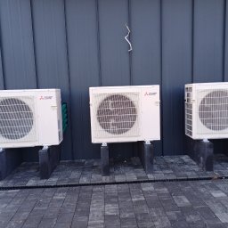 MAT-CAR Mateusz Woźniak - Trzy jednostki zewnętrzne klimatyzacji Mitsubishi Electric zamontowane na betonowych podstawach przy ciemnej elewacji budynku. Widoczne przewody i kostka brukowa.