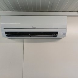 MAT-CAR Mateusz Woźniak - Biały klimatyzator Mitsubishi Electric zamontowany na jasnej ścianie w Mniowie. Widoczne detale urządzenia i fragment sufitu. Minimalistyczny design.