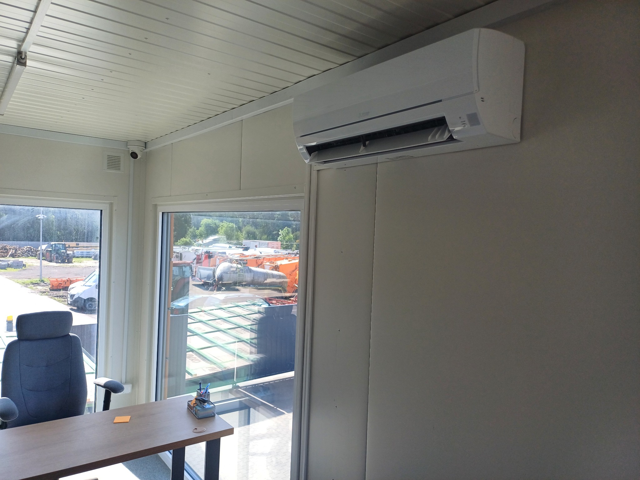 Biały klimatyzator ścienny w jasnym pomieszczeniu biurowym z widokiem na plac budowy przez okno. Widoczny stół i krzesło biurowe.