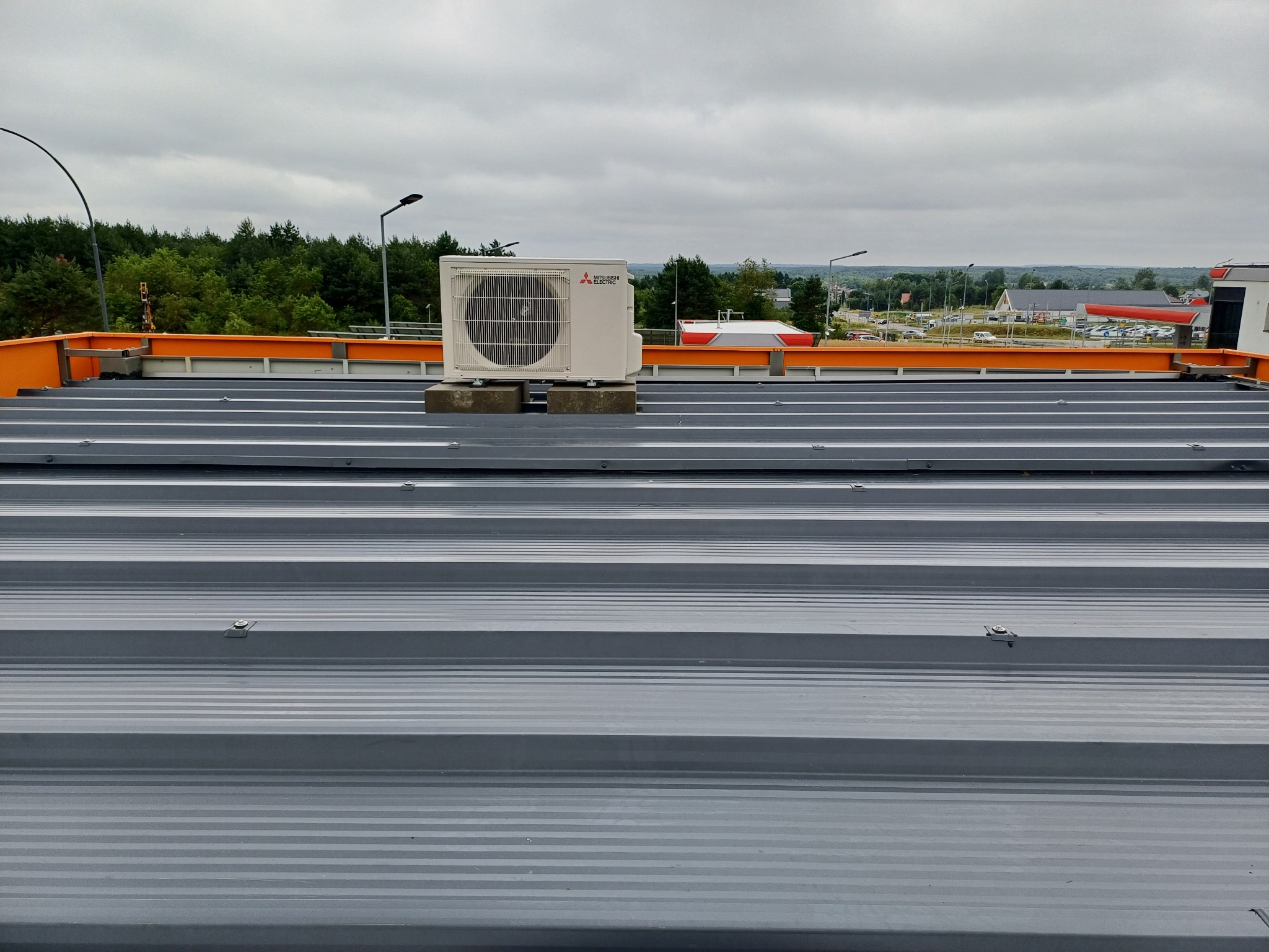 Biała jednostka zewnętrzna klimatyzacji Mitsubishi Electric na szarym, blaszanym dachu z pomarańczową barierką. W tle drzewa i pochmurne niebo.
