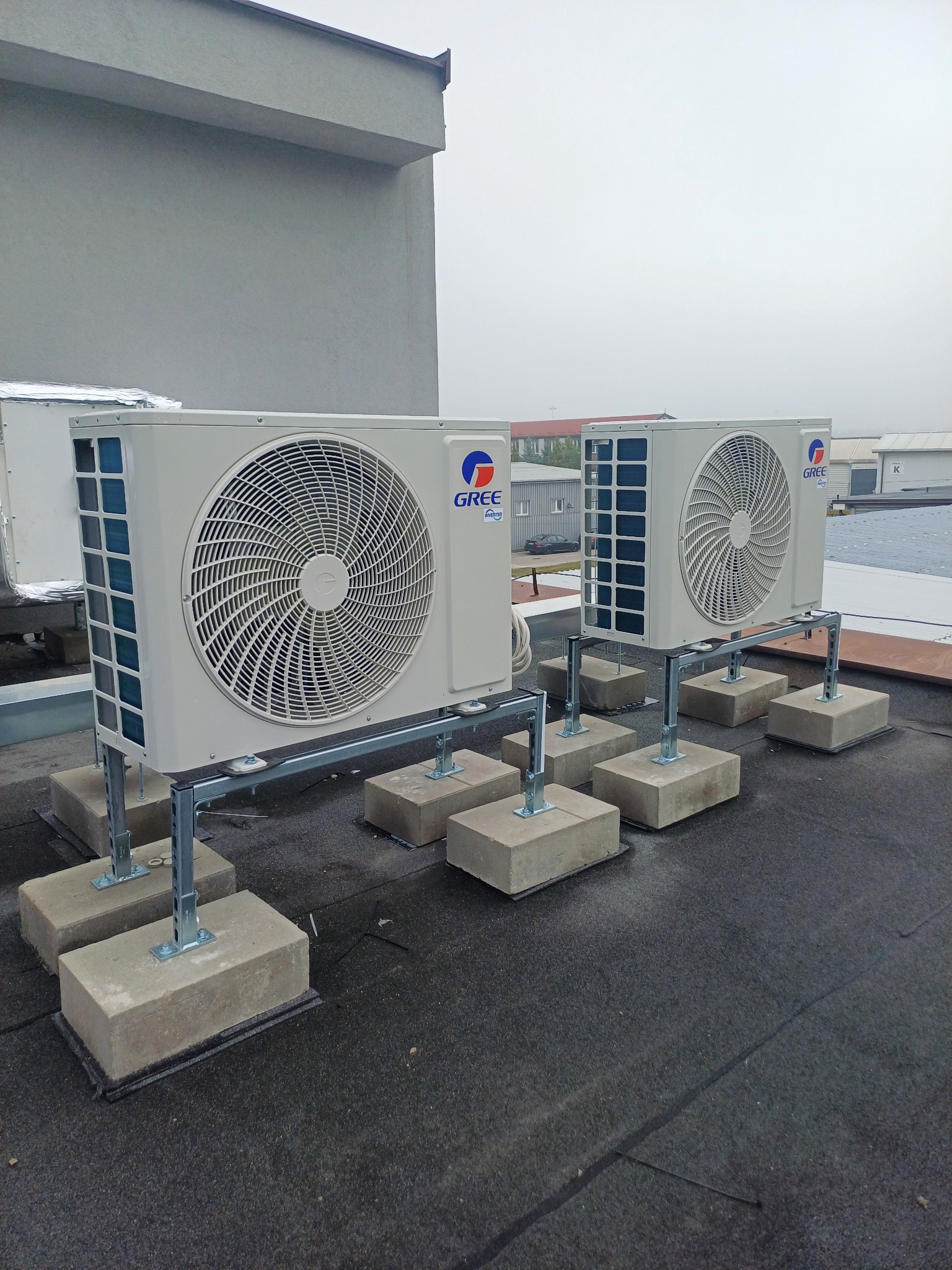 Dwa klimatyzatory Gree zamontowane na dachu budynku, na betonowych podstawach z metalowymi wspornikami. Widoczne budynki w tle i pochmurne niebo.