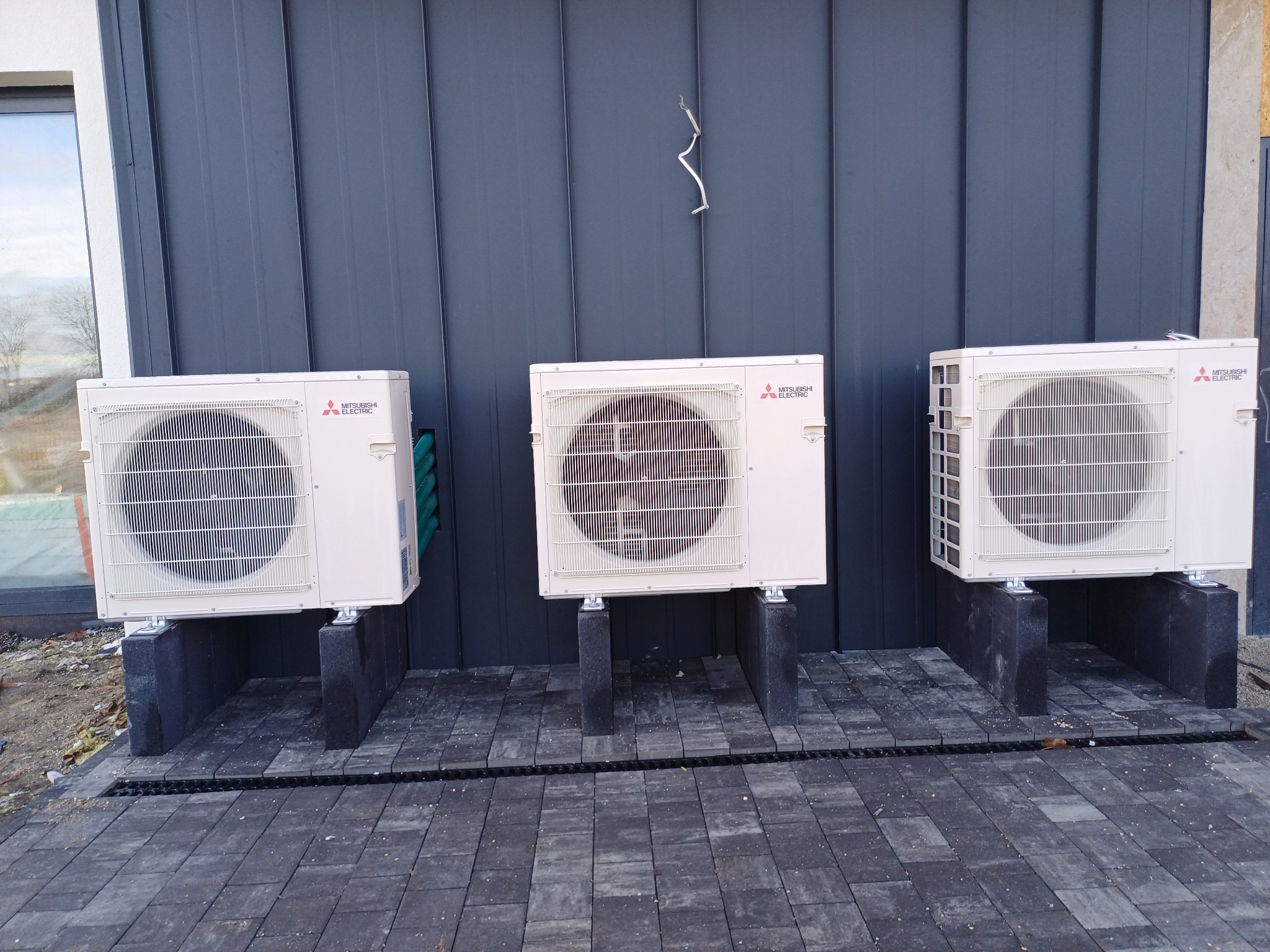 Trzy jednostki zewnętrzne klimatyzacji Mitsubishi Electric zamontowane na betonowych podstawach przy ciemnej elewacji budynku. Widoczne przewody i kostka brukowa.