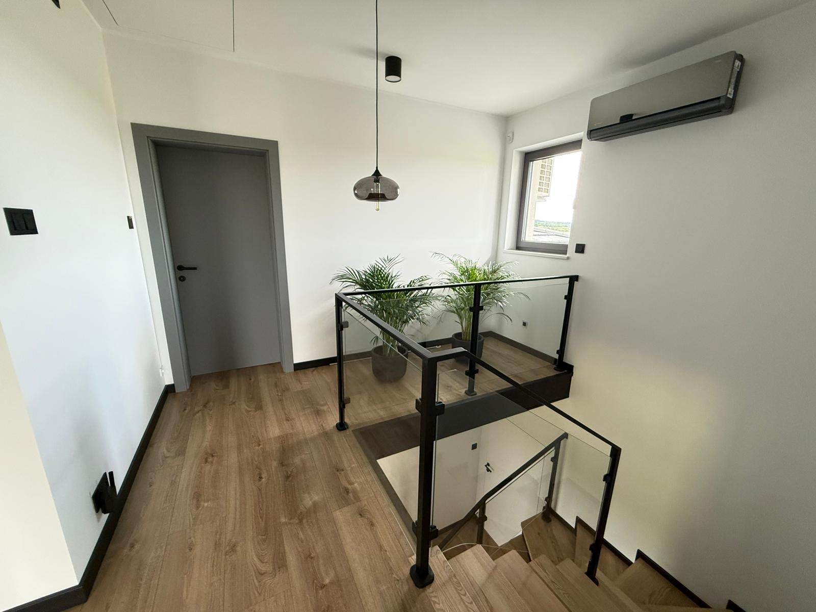 Nowoczesne wnętrze domu: drewniane schody z czarną balustradą i szklanymi panelami, parkiet, szara stolarka drzwiowa, klimatyzator i rośliny doniczkowe.
