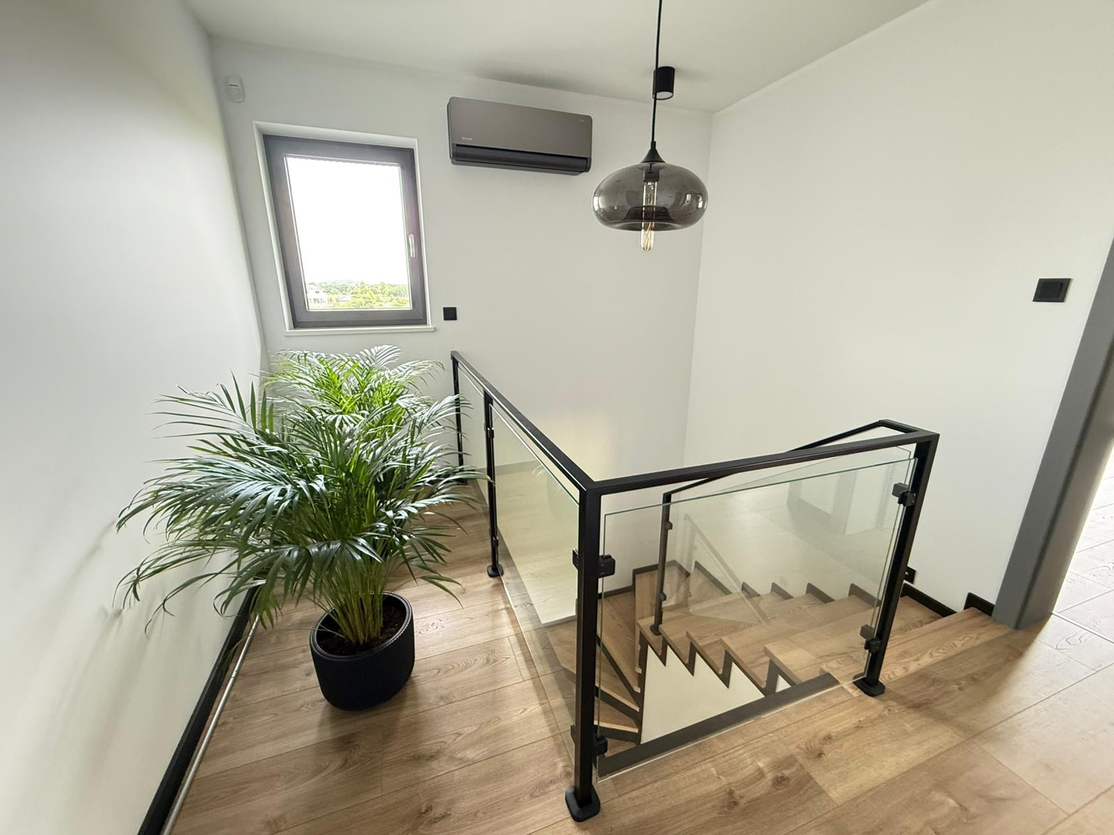 Nowoczesne schody z drewnianymi stopniami i szklaną balustradą, oświetlone wiszącą lampą. Na podłodze parkiet, obok donica z zieloną rośliną. Widoczny klimatyzator na ścianie.
