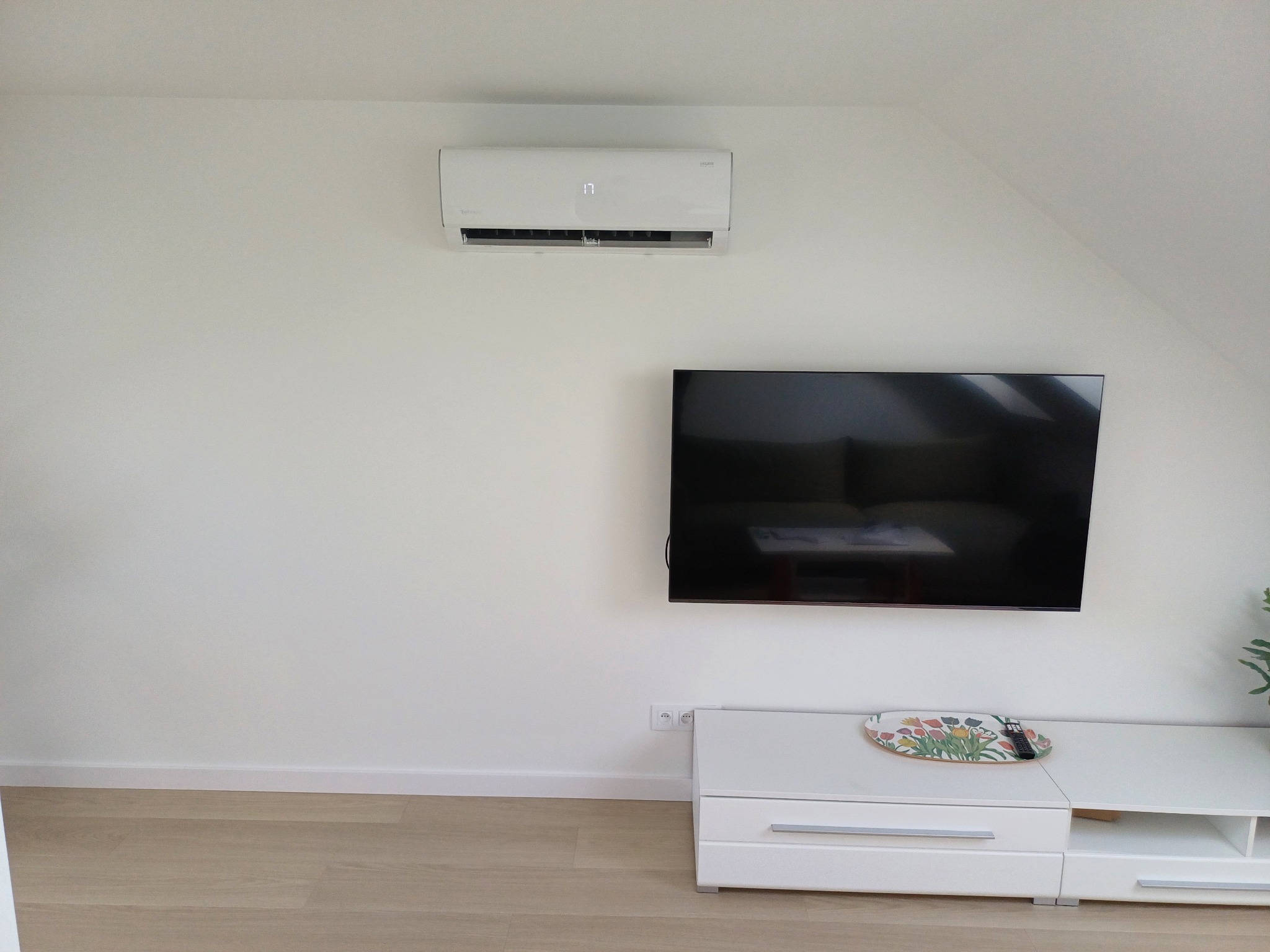 Biała klimatyzacja ścienna w jasnym pokoju z telewizorem i białą komodą. Minimalistyczny design, skandynawski styl. Widoczny fragment rośliny doniczkowej.