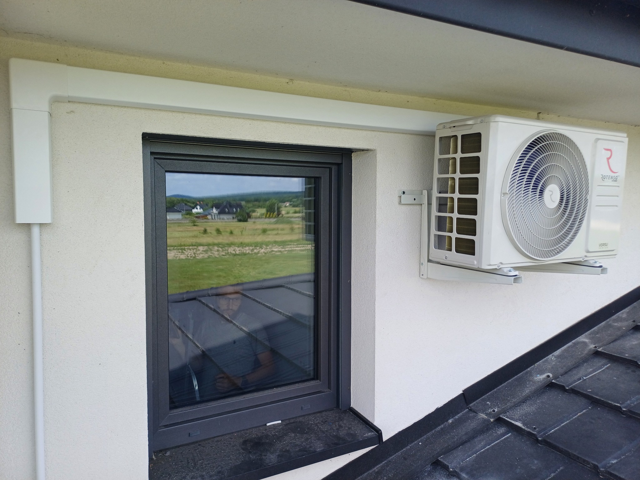 Biały klimatyzator Rotenso zamontowany na elewacji obok okna z ciemną ramą. Widoczne elementy instalacji i fragment dachu. W oknie odbija się osoba.
