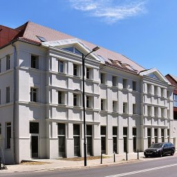 GAP NIERUCHOMOŚCI SPÓŁKA Z OGRANICZONĄ ODPOWIEDZIALNOŚCIĄ - Odnowiony budynek biurowy z jasną elewacją, czerwoną nadbudówką i samochodami zaparkowanymi wzdłuż ulicy w słoneczny dzień. Widok z perspektywy ulicy.