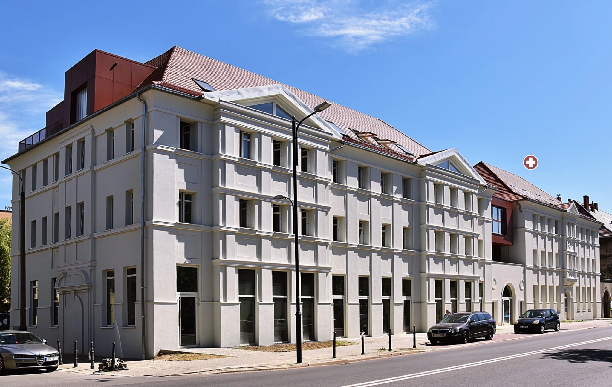 Odnowiony budynek biurowy z jasną elewacją, czerwoną nadbudówką i samochodami zaparkowanymi wzdłuż ulicy w słoneczny dzień. Widok z perspektywy ulicy.