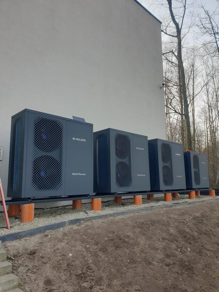 Szereg szarych pomp ciepła MultiTherma Igloo ustawionych na pomarańczowych wspornikach przed budynkiem z szarą elewacją i drzewami w tle.