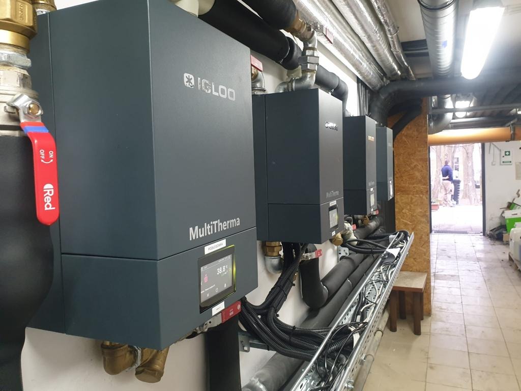 Rząd trzech szarych kotłów grzewczych MultiTherma z logo Igloo zamontowanych na ścianie w kotłowni, z widocznymi rurami, przewodami i zaworem Red, otwartymi drzwiami na zewnątrz w tle.